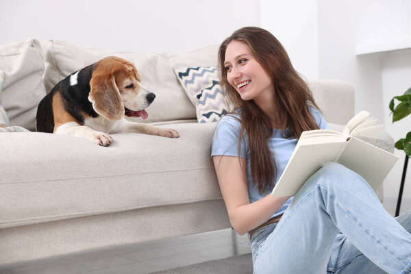 Happy young woman reading book near her cute Beagle dog on couch at home. Lovely pet