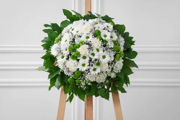 Funeral wreath of flowers on wooden stand near white wall indoors