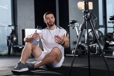 Man with bottle of water streaming online training on phone at gym. Fitness coach