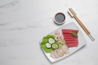 Sashimi seti (çiğ ton balığı, yağlı balık ve karides dilimleri) beyaz mermer masa üzerinde salatalık, marul, vasabi ve soya sosuyla servis edilir. Metin için boşluk