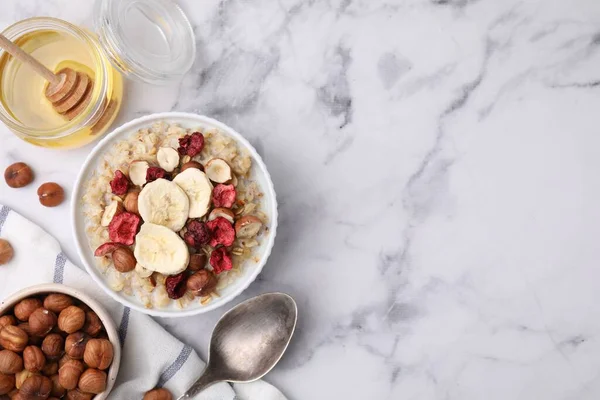 stock image Delicious oatmeal with freeze dried berries, banana and hazelnuts served on white marble table, flat lay. Space for text