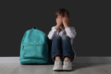 Siyah duvarın yanında sırt çantalı üzgün bir çocuk oturuyor. Okul zorbalığı