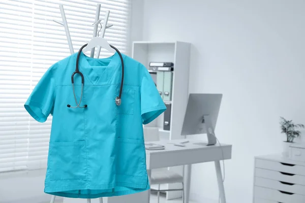 Stock image Turquoise medical uniform and stethoscope hanging on rack in clinic. Space for text