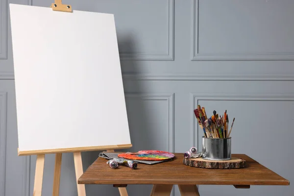 stock image Easel with blank canvas and different art supplies on wooden table near grey wall