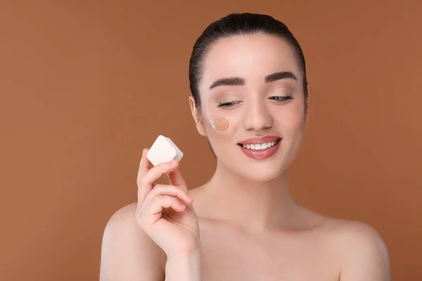 Woman applying foundation on face with makeup sponge against brown background