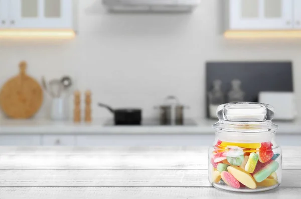 Glass jar with tasty gummy candies on white wooden table in kitchen. Space for text