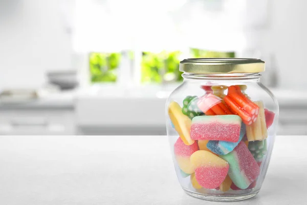 Glass jar with tasty gummy candies on white table in kitchen. Space for text