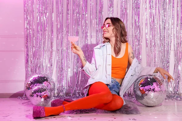Beautiful woman in stylish outfit with glass of drink among disco balls at party indoors