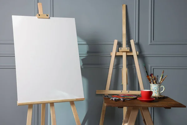 stock image Easel with blank canvas, cup of drink and different art supplies on wooden table near grey wall