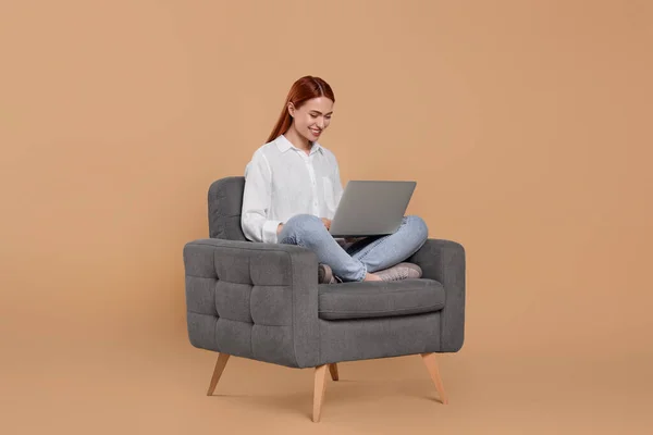 在米色背景的扶手椅上与笔记本电脑一起工作的年轻女性面带微笑 — 图库照片