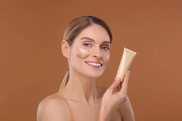 Woman holding tube of foundation on brown background