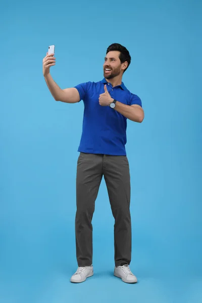 Uśmiechnięty Mężczyzna Robi Sobie Selfie Pomocą Smartfona Pokazuje Kciuki Jasnoniebieskim — Zdjęcie stockowe