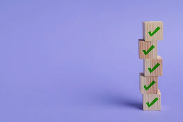 Stacked wooden cubes with check marks on light violet background. Space for text