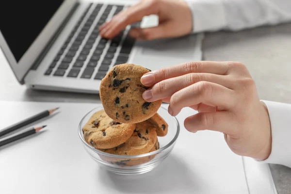 女性はオフィスでラップトップで作業中にボウルからチョコレートチップクッキーを取ります クローズアップ — ストック写真