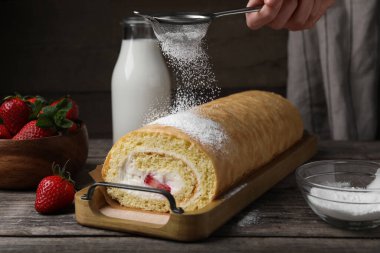 Kadın, ahşap masada çilek ve kremayla lezzetli bir kek rulosunun üzerine pudra şekeri döküyor.