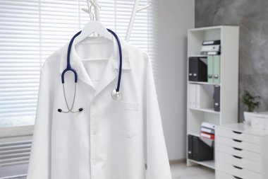 Klinikte askıda asılı duran beyaz doktor önlüğü ve steteskop, yakın plan. Metin için boşluk