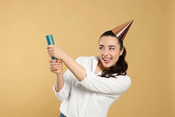 Young woman blowing up party popper on light brown background