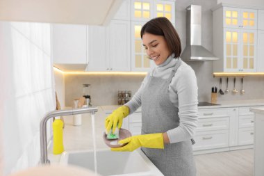 Modern mutfakta lavabonun üstünde genç ve mutlu bir kadın bulaşık yıkıyor.