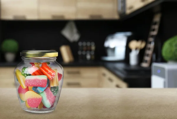 Glass jar with tasty gummy candies on light brown table in kitchen. Space for text