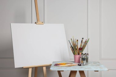 Easel with blank canvas and different art supplies on wooden table near white wall clipart