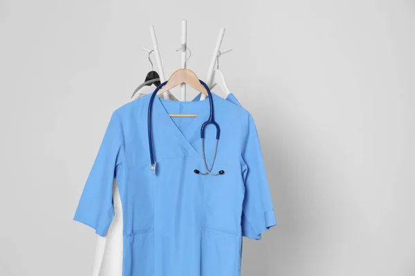 Medical uniforms and stethoscope hanging on rack against light grey background. Space for text