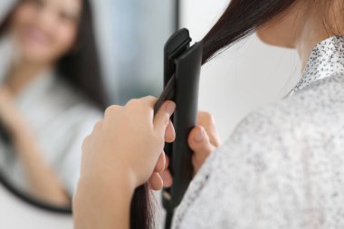 Woman using hair iron near mirror indoors, closeup. Space for text