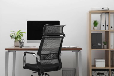 Cozy workspace with computer on desk, chair and bookcase near white wall at home