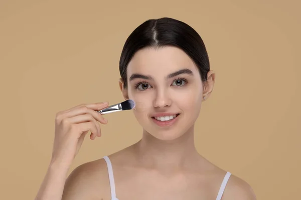 Teenage girl applying foundation on face with brush against beige background