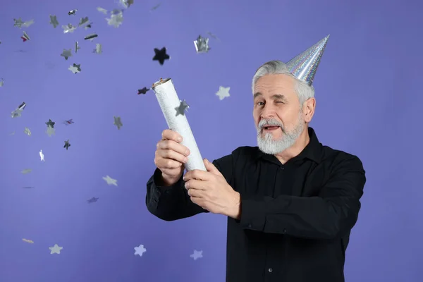 Man blowing up party popper and winking on purple background