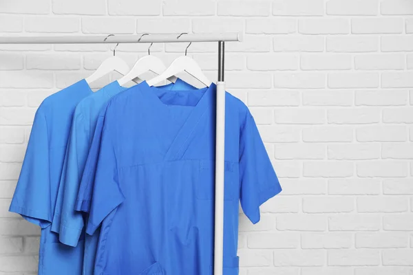 Medical uniforms hanging on rack near white brick wall. Space for text