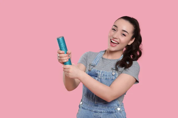 Young woman blowing up party popper on pink background, space for text