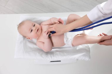 Pediyatrist, klinikte steteskopla küçük şirin bir bebeği tartıyor ve muayene ediyor.