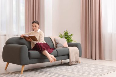 Kadın pencerenin yanındaki kanepede kitap okuyor ve evinde şık perdeler var. Metin için boşluk