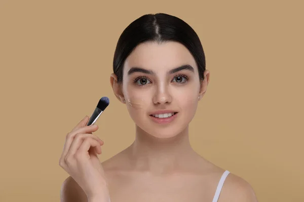 Teenage girl applying foundation on face with brush against beige background