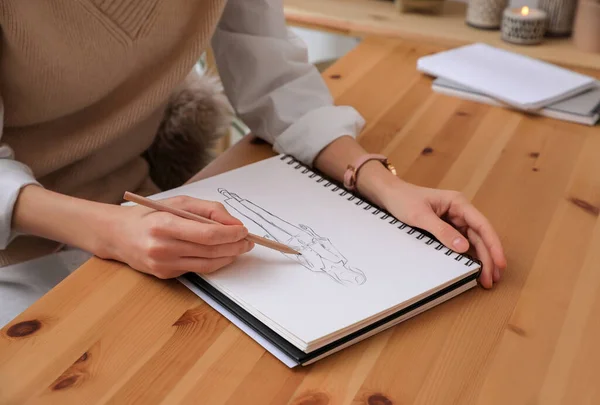 Fashion designer creating new look. Woman drawing sketch in book with pencil at wooden table, closeup