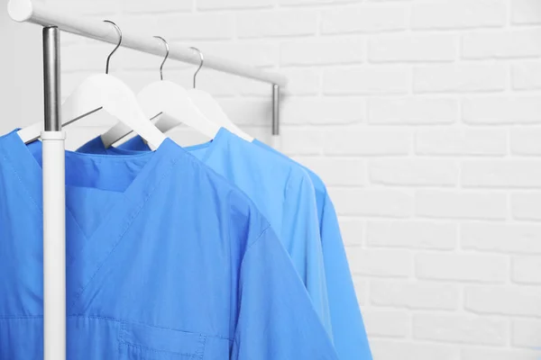 Medical uniforms hanging on rack near white brick wall. Space for text