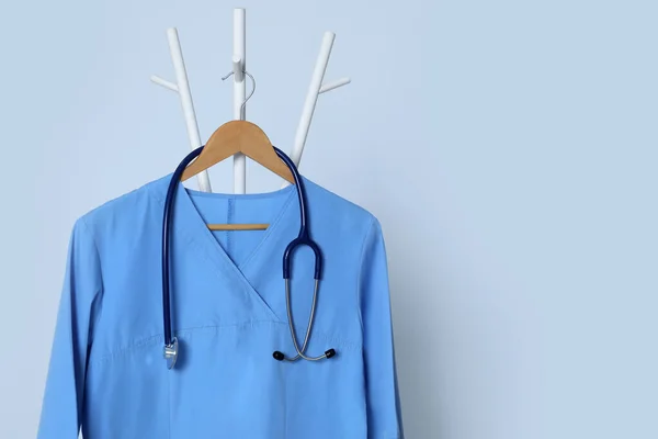 Medical uniform and stethoscope hanging on rack against light grey background. Space for text