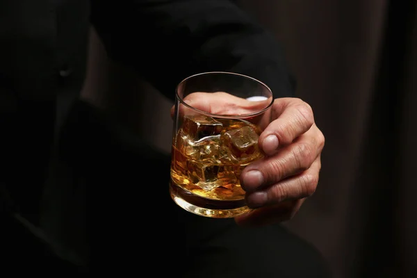 stock image Man holding glass of whiskey with ice cubes on dark background, closeup