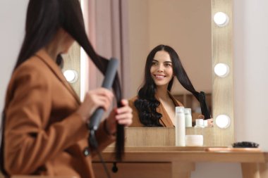 Güzel, mutlu bir kadın odada aynanın yanında saç maşası kullanıyor.