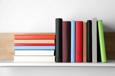 Beyaz duvarın yanındaki ahşap rafta bir sürü ciltli kitap var.