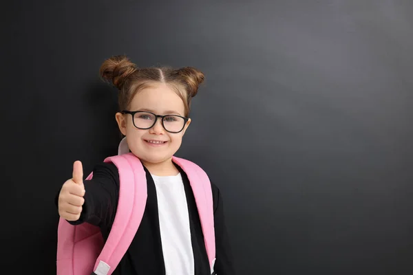 Happy little school child with backpack showing thumbs up near chalkboard. Space for text