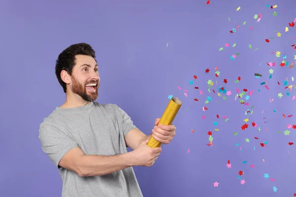 Emotional man blowing up party popper on violet background