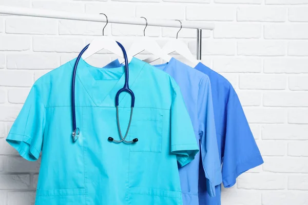 Medical uniforms and stethoscope hanging on rack near white brick wall
