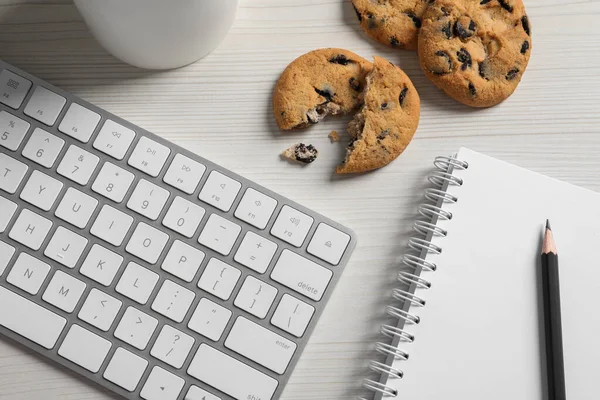 Chocolate chip cookies, keyboard and office supplies on white wooden table, flat lay