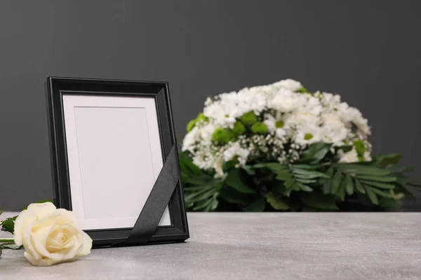 Photo frame with black ribbon, rose on light table and wreath of flowers near grey wall indoors. Funeral attributes