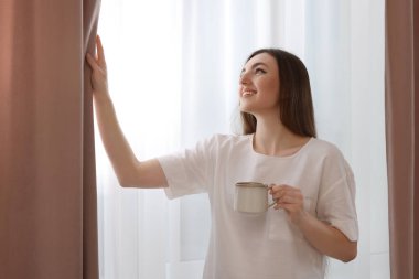 Bir fincan sıcak içecekle evde şık perdeler açan bir kadın.