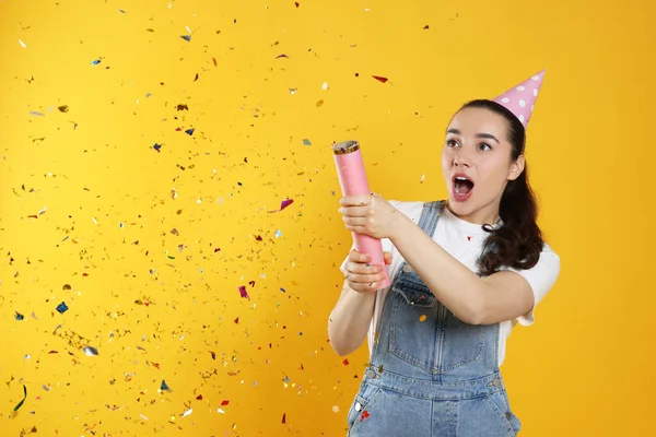 Young woman blowing up party popper on yellow background