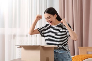 Emotional young woman opening parcel at home. Internet shopping clipart