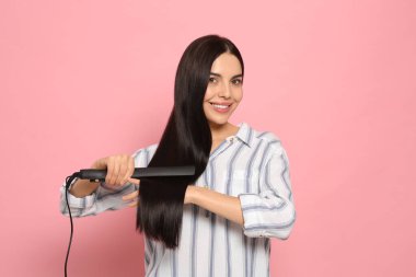 Güzel, mutlu kadın pembe arka planda saç ütüsü kullanıyor.