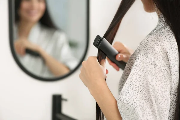 Woman using hair iron near mirror indoors, closeup. Space for text